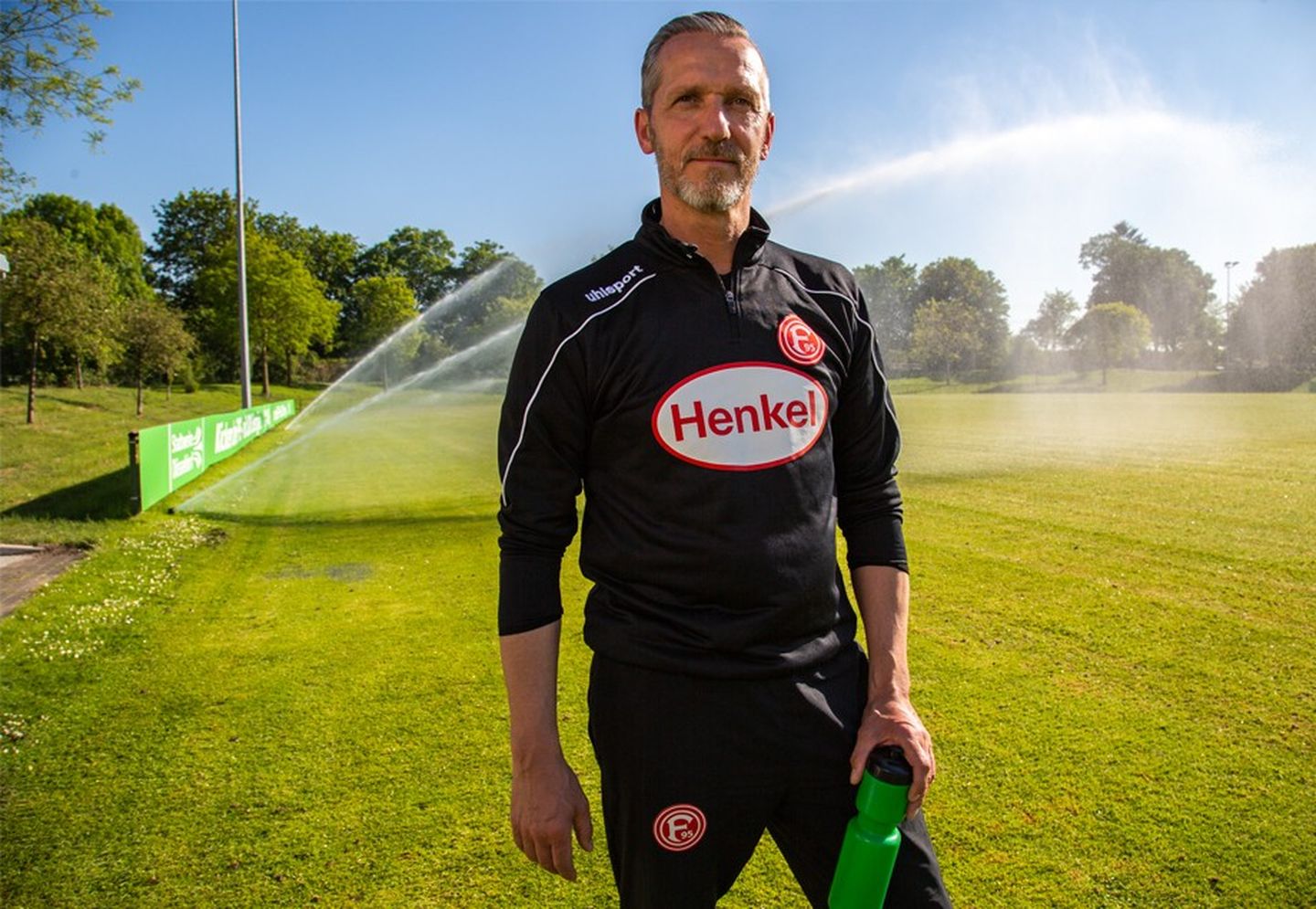 Carsten Fiedler steht mit einer Trinkflasche in der Hand auf dem Trainingsgelände der Fortuna vor einem laufenden Rasensprenger.
