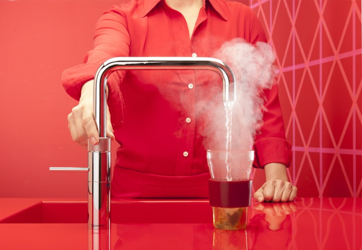 Eine Frau in Rot, die einen smarten Wasserhahn in einer roten Küche bedient, um kochendes Wasser in eine Teetasse laufen zu lassen.