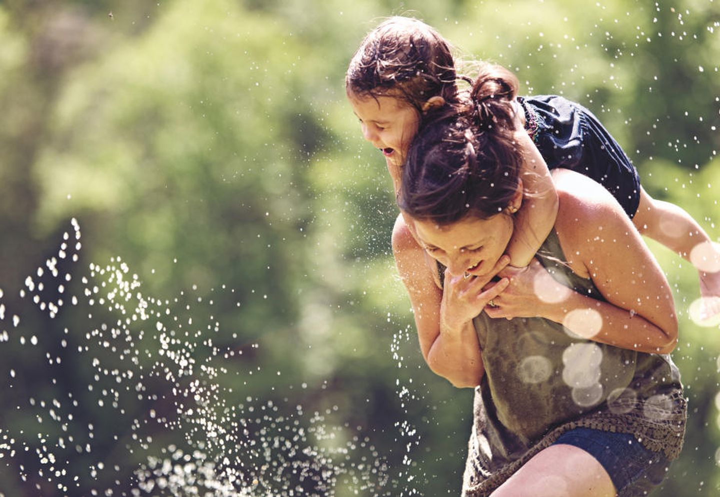 Nahaufnahme von Mutter und Kind, die auf einem Wasserspielplatz durch eine Wasserfontäne laufen. © lisegagne / E+ via Getty Images