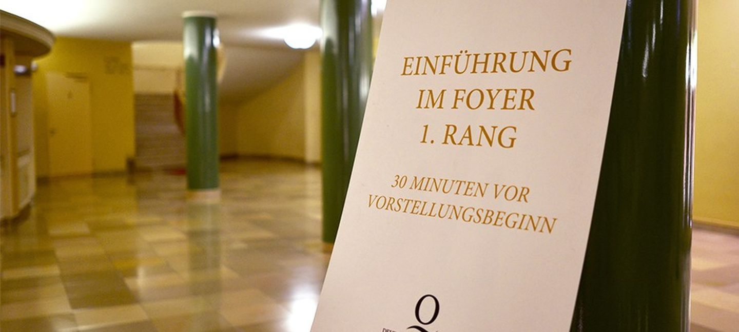 Entree einer Oper mit hellem Marmorboden und grünen Säulen, im Vordergrund eine Staffelei mit einem Plakat, auf dem steht: Einführung im Foyer. 30 Minuten vor Vorstellungsbeginn.