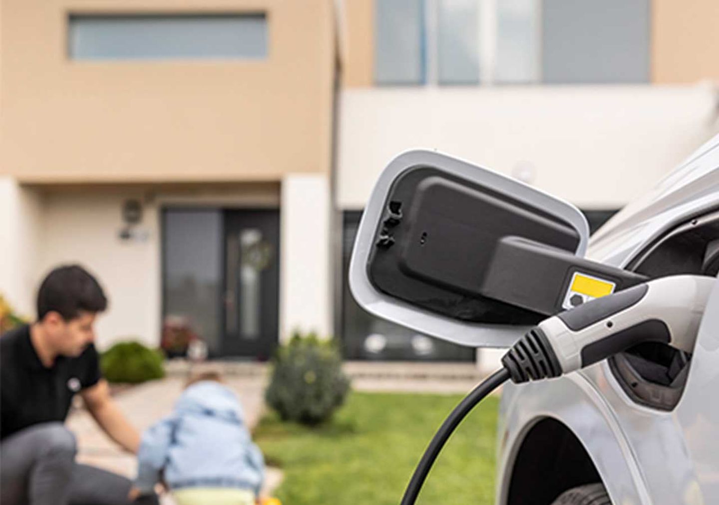 Ein E-Auto wird zuhause geladen. Im Hintergrund: Ein Vater mit seinem Kind.