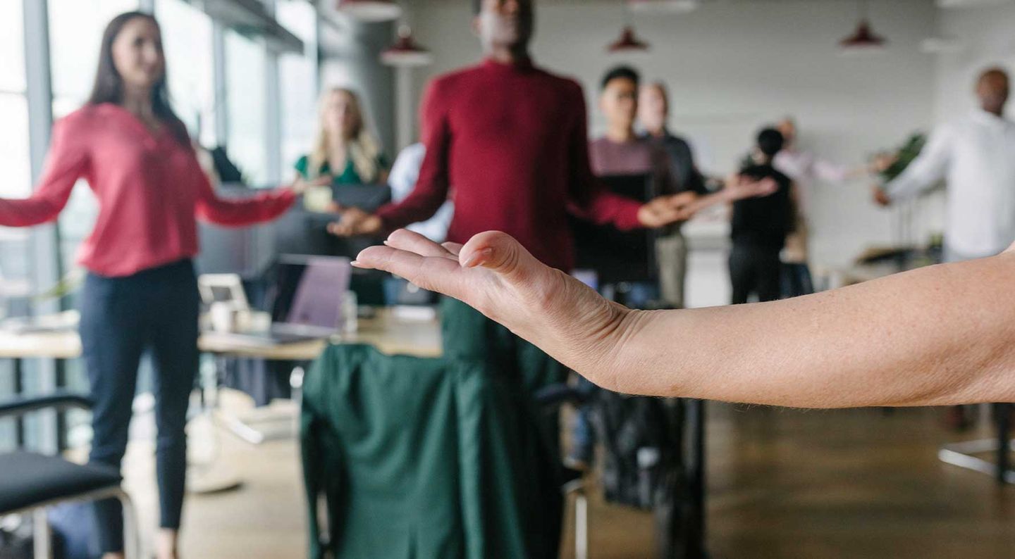 Kolleg:innen machen an ihren Tischen im Büro eine Übung im Stehen, indem sie die Arme seitlich halten und den Rücken dehnen. © Hinterhaus Productions / DigitalVision via Getty Images