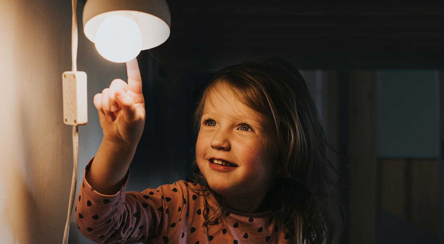 Ein kleines Mädchen sitzt vor einer eingeschalteten Lampe und berührt die Glühbirne mit dem Finger. © Catherine Falls Commercial / Moment via Getty Images