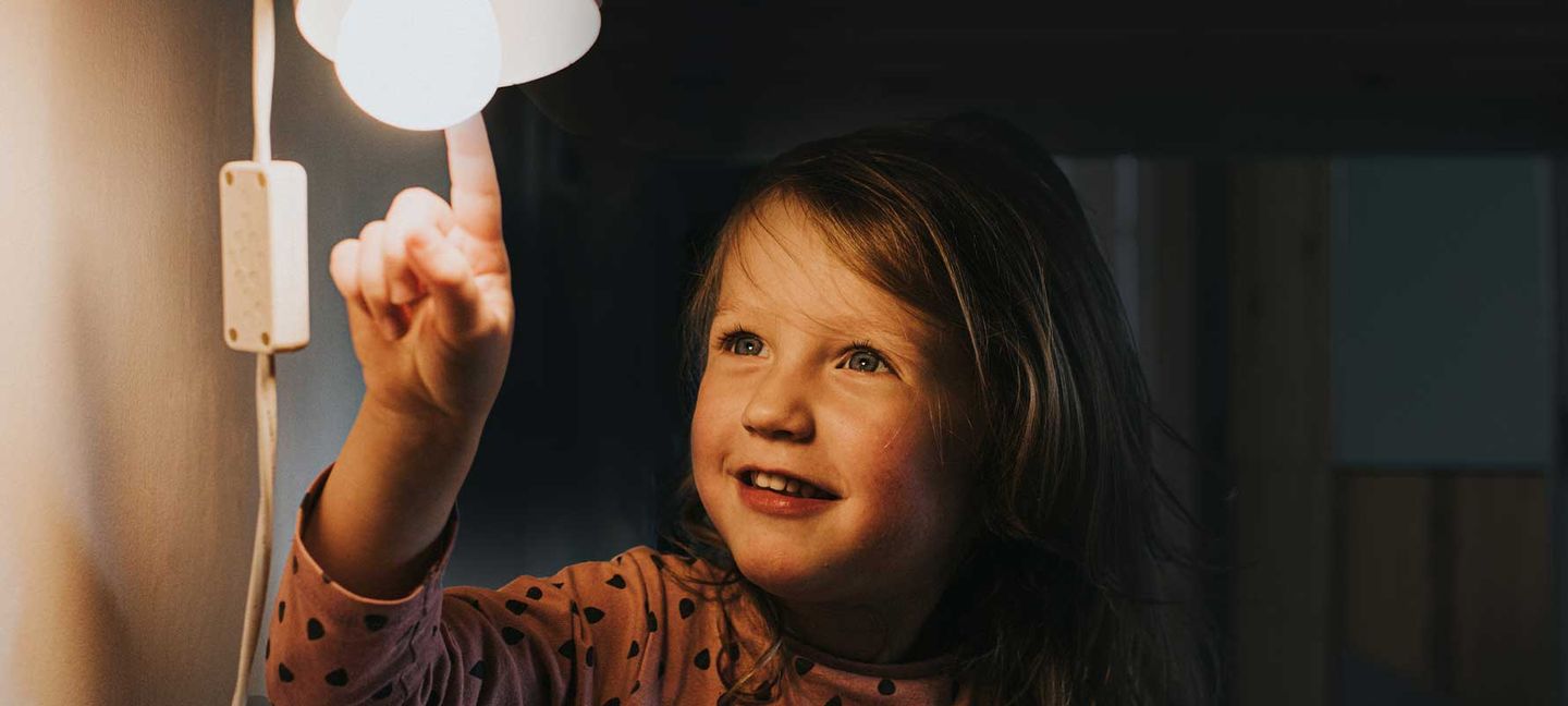 Ein kleines Mädchen sitzt vor einer eingeschalteten Lampe und berührt die Glühbirne mit dem Finger. © Catherine Falls Commercial / Moment via Getty Images