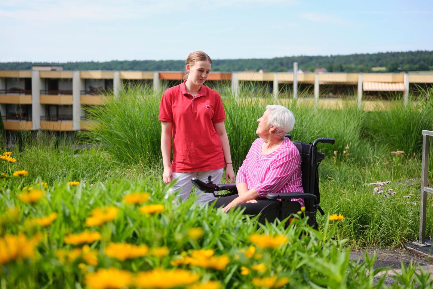 Junge Pflegerin spricht mit Seniorin im Rollstuhl in einem begrünten Außenbereich. © AWO Bundesverband © AWO Bundesverband
