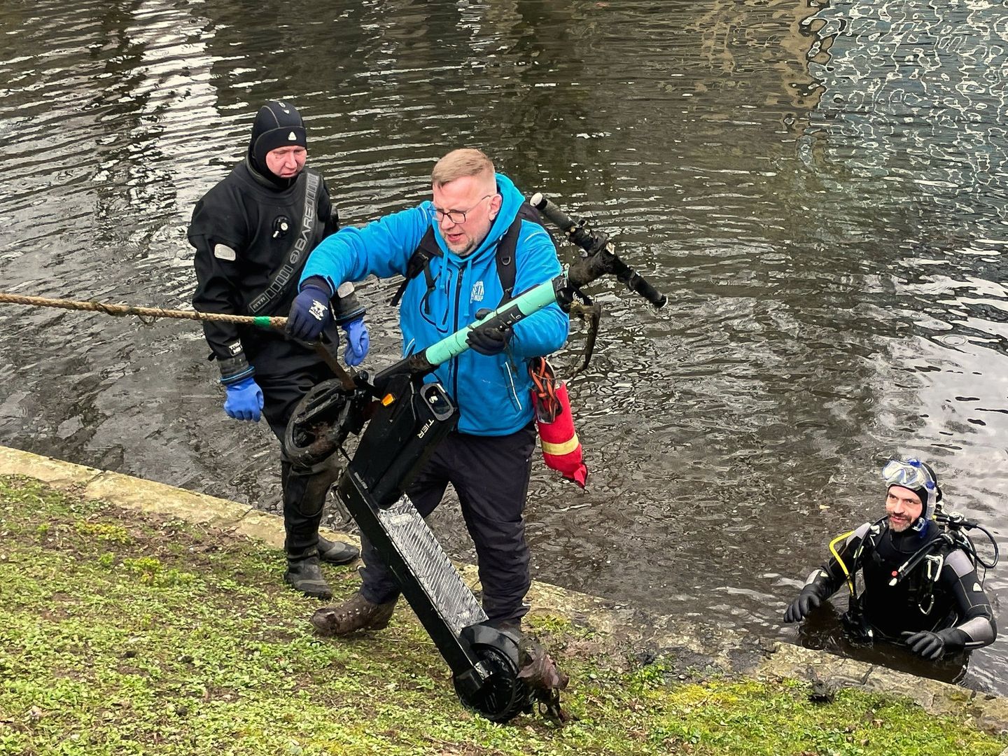 Zwei Taucher und ein Helfer bergen einen E-Scooter aus einem Wassergraben. © Pro Düsseldorf e. V.