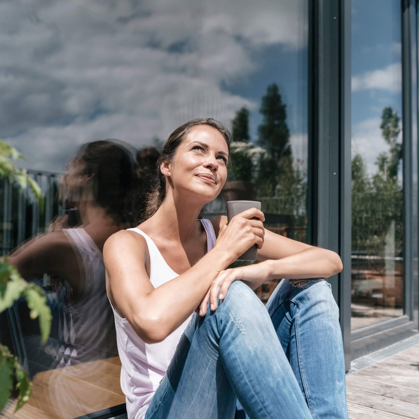 Eine Frau sitzt auf der Terrasse ihres Hauses in der Sonne und hält ein Getränk in der Hand. © Westend61 via Getty Images