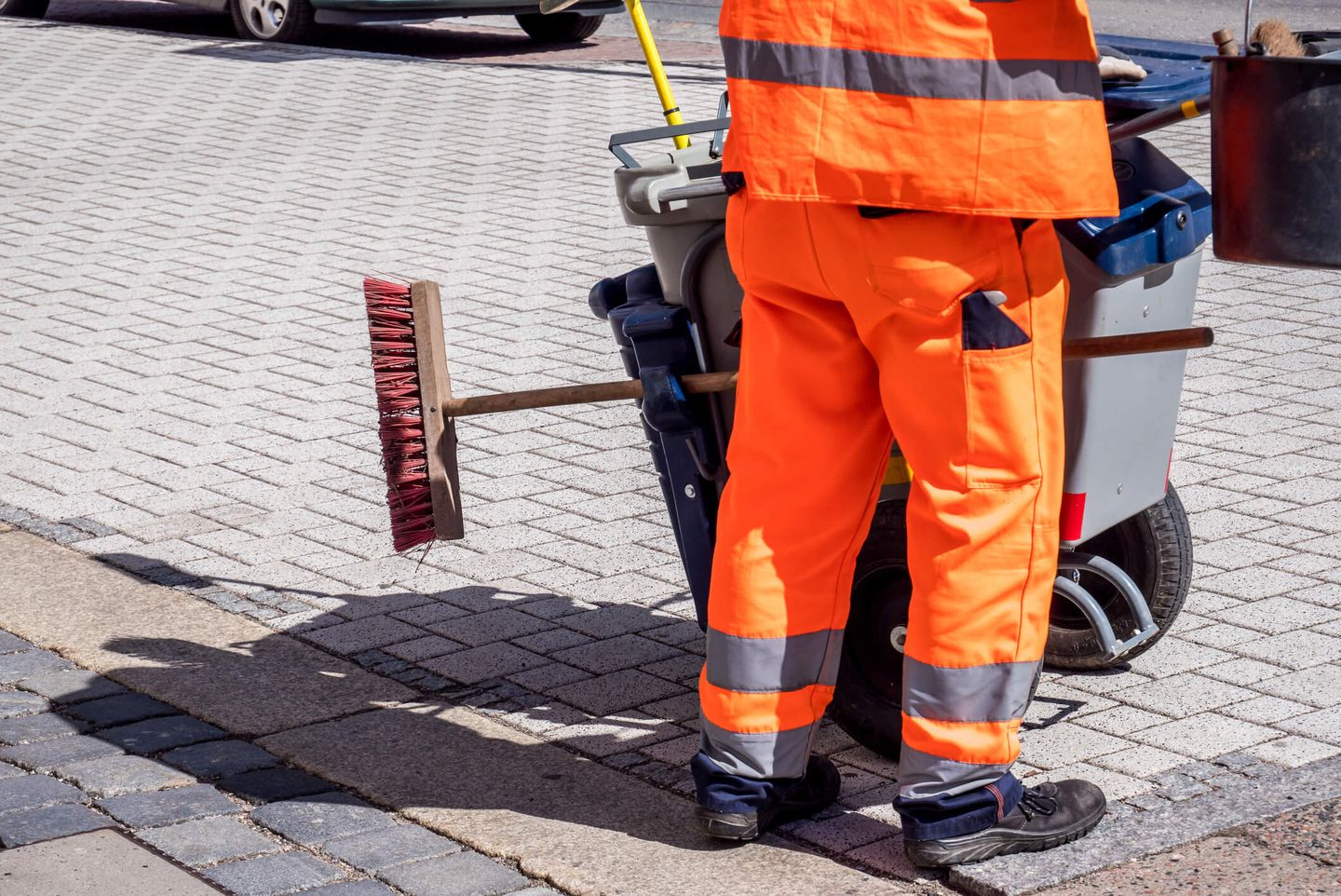 Eine Reinigungsfachkraft steht in orangem Arbeitsanzug an einem Putzwagen, an dem längs ein Besen befestigt ist. © Animaflora / iStock / Getty Images Plus via Getty Images © Animaflora / iStock / Getty Images Plus via Getty Images