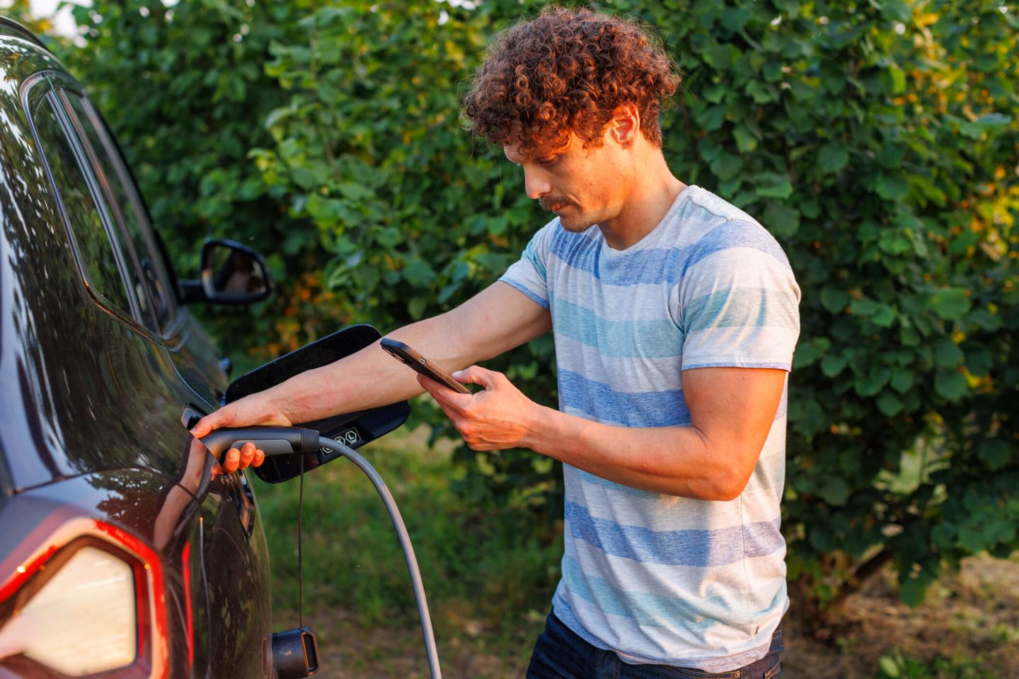 Ein Mann steht mit seinem Smartphone in der Hand an seinem E-Auto, das aufgeladen wird. © vgajic / E+ via Getty Images
