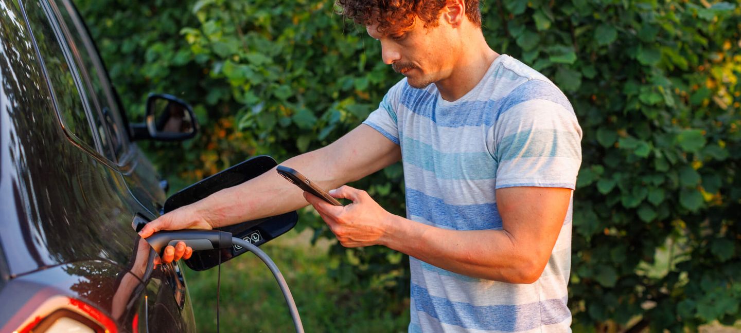 Ein Mann steht mit seinem Smartphone in der Hand an seinem E-Auto, das aufgeladen wird. © vgajic / E+ via Getty Images