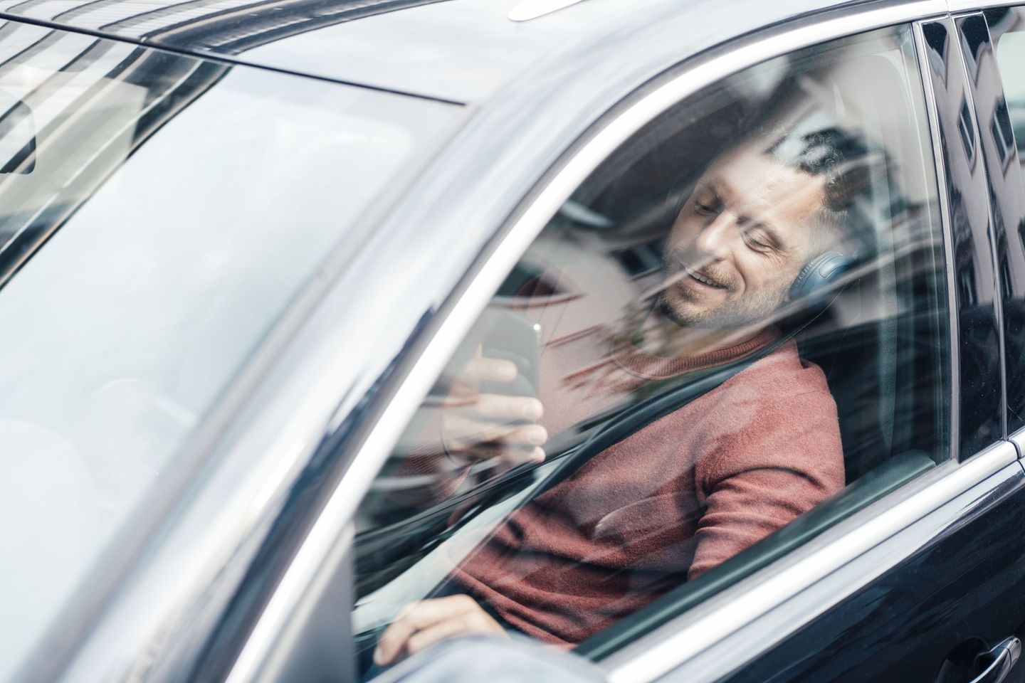 Ein Mann sitzt lächelnd hinterm Steuer und schaut auf sein Smartphone.  © Westend61 / Westend61 via Getty Images