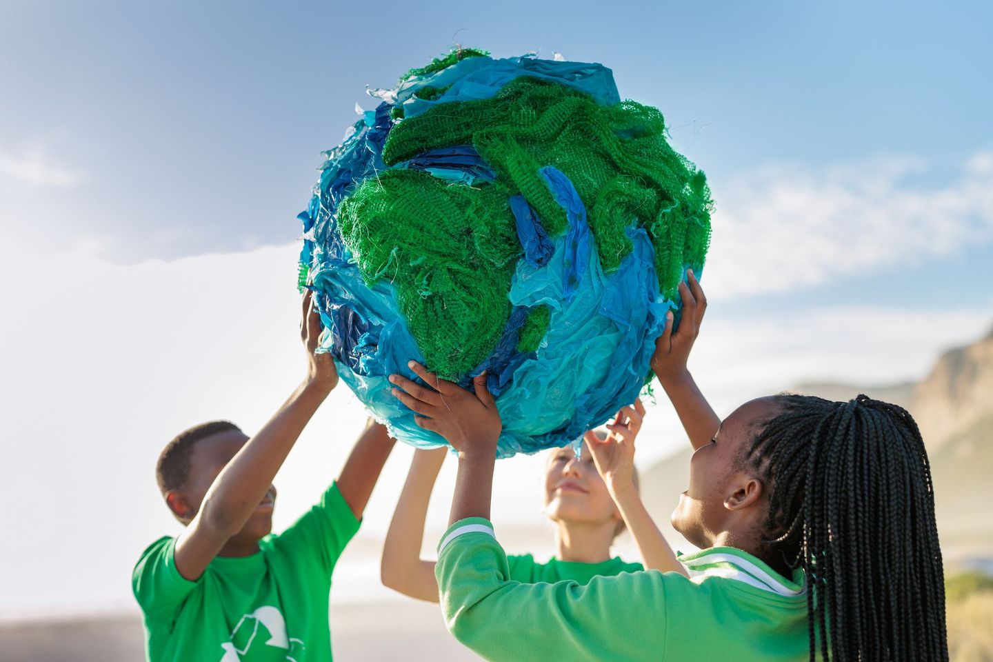 Drei Kinder in grünen T-Shirts halten eine große, aus recycelten Materialien gefertigte Weltkugel hoch. © Alistair Berg / DigitalVision via Getty Images