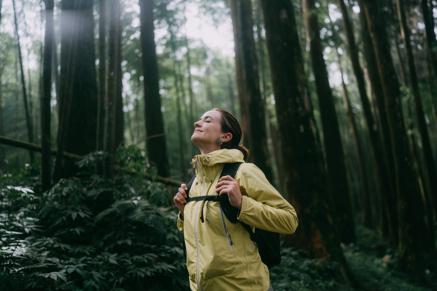 Eine Frau steht mit Regenjacke und Rucksack im Wald und atmet mit geschlossenen Augen tief ein. © Ippei Naoi / Moment via Getty Images © Ippei Naoi / Moment via Getty Images