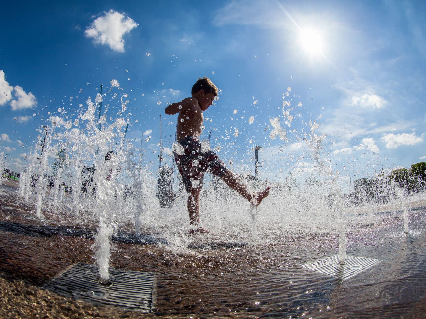 Kind spielt in Wasserfontänen auf einem Platz bei strahlendem Sommerwetter. © paul mansfield photography / Moment via Getty Images