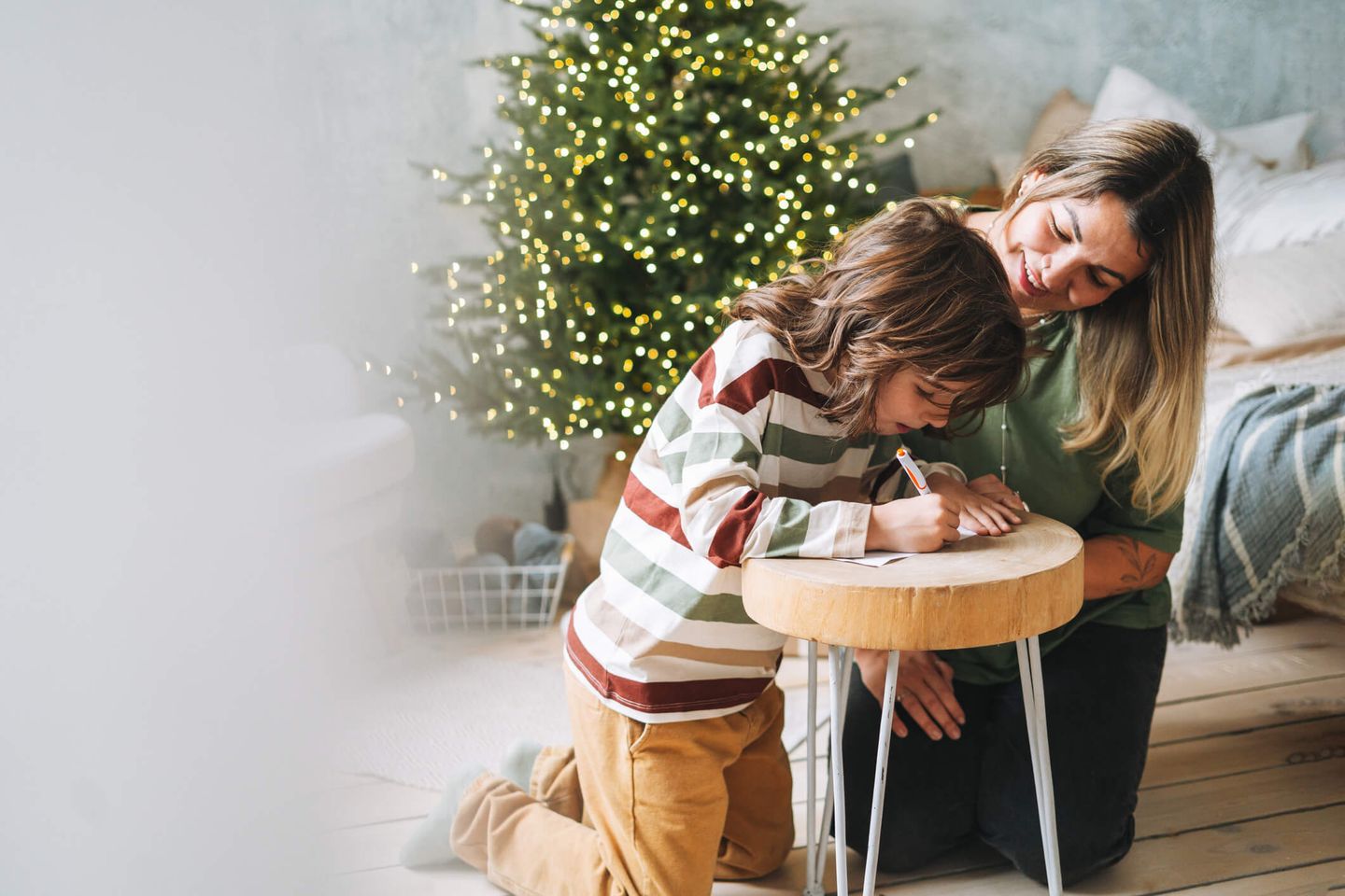 Eine Mutter und ihr Kind hocken auf dem Boden und schreiben einen Wunschzettel, im Hintergrund steht ein beleuchteter Weihnachtsbaum. © Galina Zhigalova / Moment via Getty Images © Galina Zhigalova / Moment via Getty Images