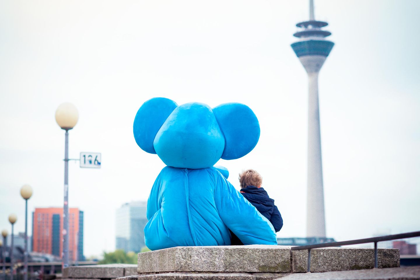 Das Maskottchen im Kostüm eines blauen Elefanten sitzt mit einem Kind auf einer Mauer und sie blicken Arm in Arm auf den Düsseldorfer Rheinturm. © Kinderschutzbund Düsseldorf