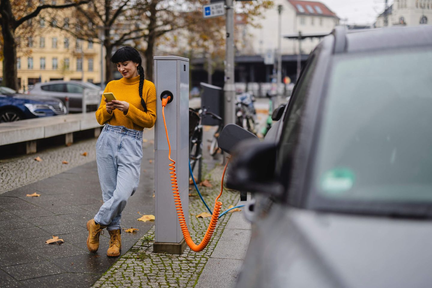 Eine junge Frau lehnt an einer Ladesäule und schaut auf ihr Handy, während ihr E-Auto geladen wird. © Westend61 via Getty Images