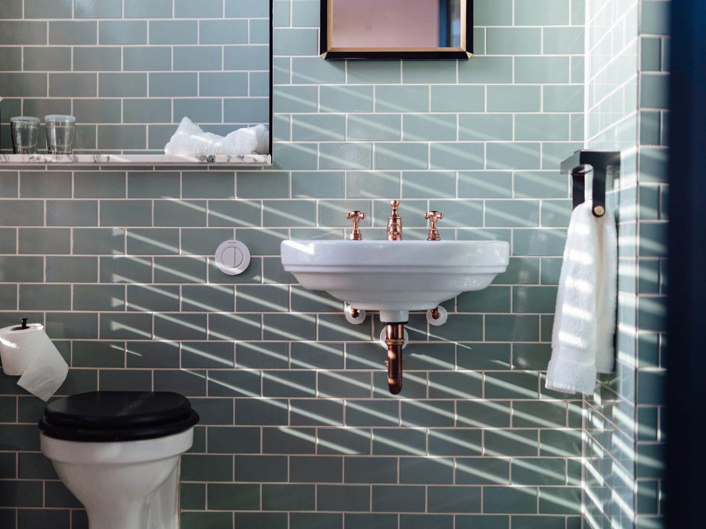 Badezimmer mit mintgrünen Metrofliesen, freihängendem weißen Waschbecken mit kupferfarbenen Armaturen, Wandregal und Toilette mit schwarzem Sitz