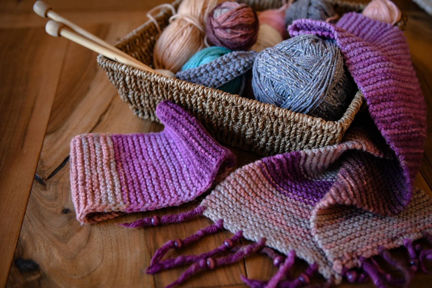 Ein geflochtener Korb mit verschiedenen Wollknäueln in Pastellfarben, Stricknadeln und einem teilweise fertig gestrickten Schal in Violett- und Rosatönen liegt auf einem Holztisch.