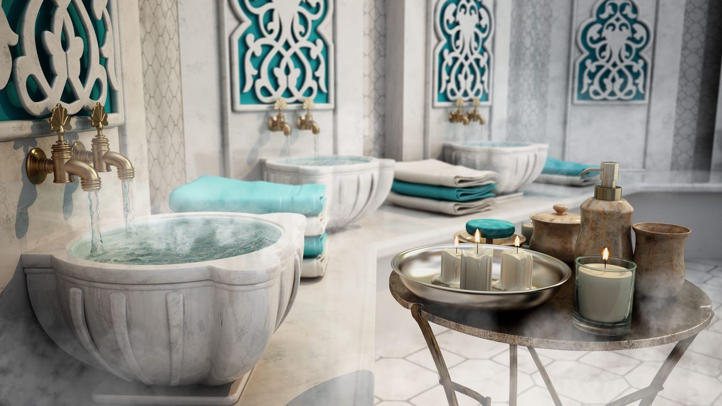 Moderne Hammam-Einrichtung mit weißen Marmorbecken, aus denen Wasser fließt, gerollten Handtüchern und dekorativen Elementen in Türkis, im Vordergrund ein Tisch mit Kerzen und Pflegeprodukten