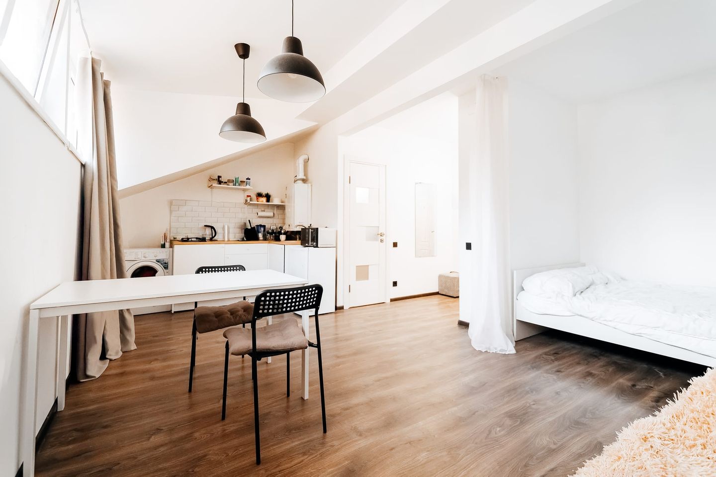 Minimalistisch eingerichtetes Studio-Apartment mit offener Küche, Esstisch mit zwei Stühlen und einem Bett in einer Ecke, durch einen Vorhang leicht abgetrennt