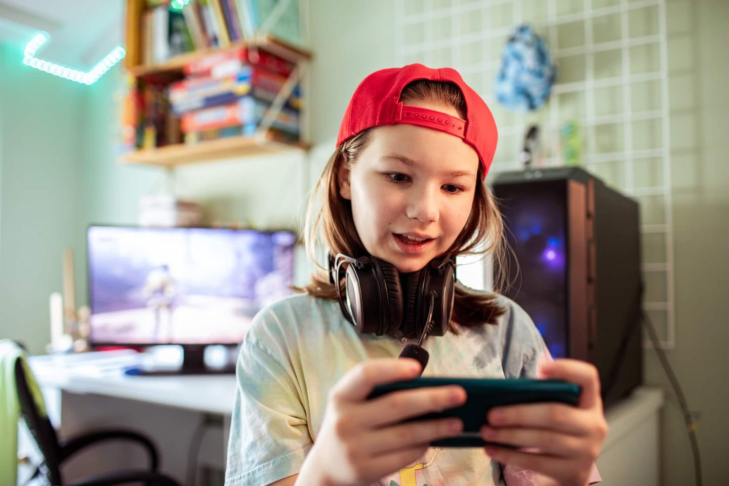 Mädchen mit roter Kappe und Kopfhörern spielt auf einem Smartphone, im Hintergrund ein Gaming-PC. Getty
