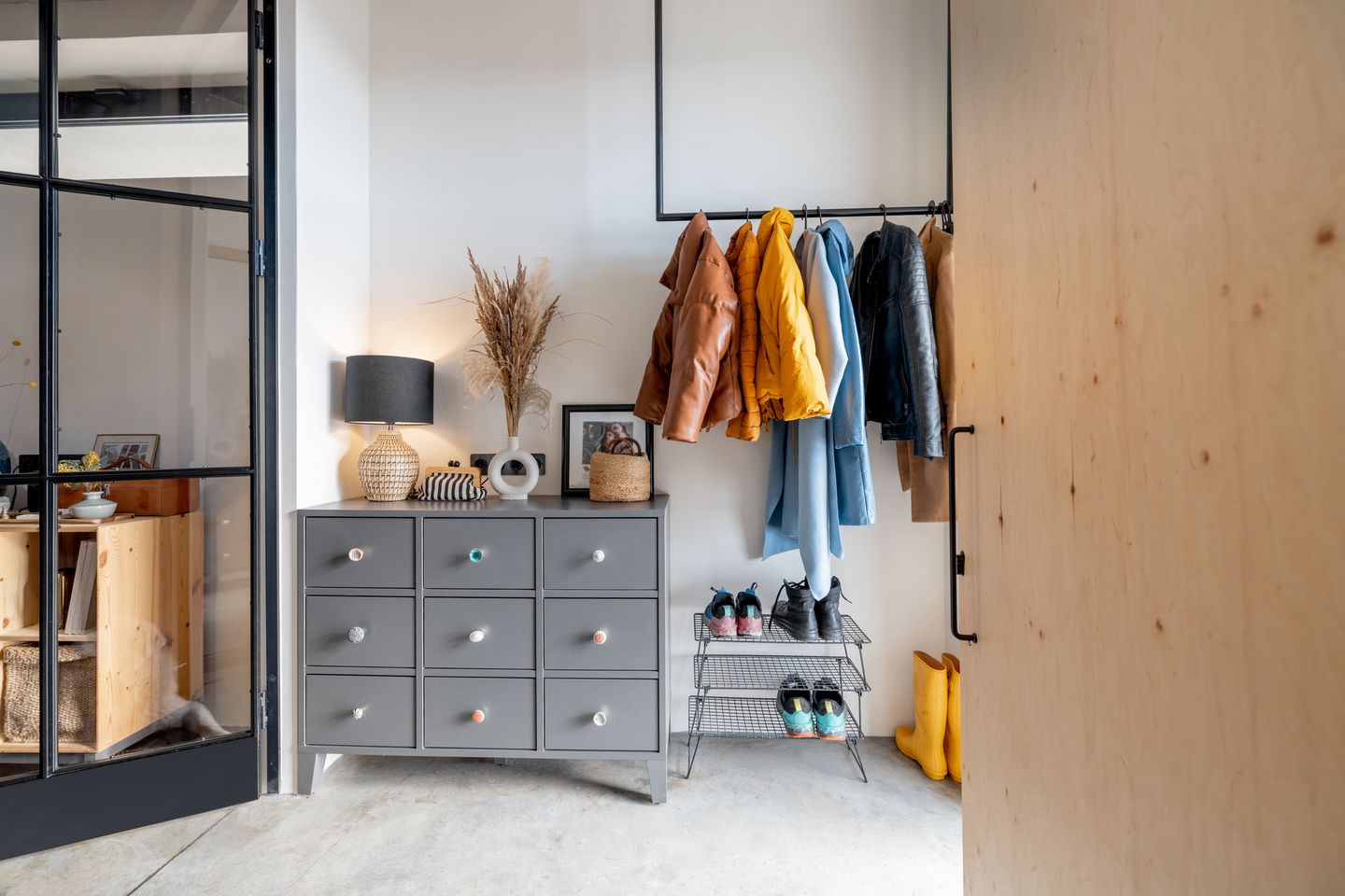 Flur mit grauer Kommode, darauf dekorative Elemente wie eine Lampe und getrocknete Gräser, darüber eine Garderobe mit bunten Jacken und Schuhen auf einem Regal.