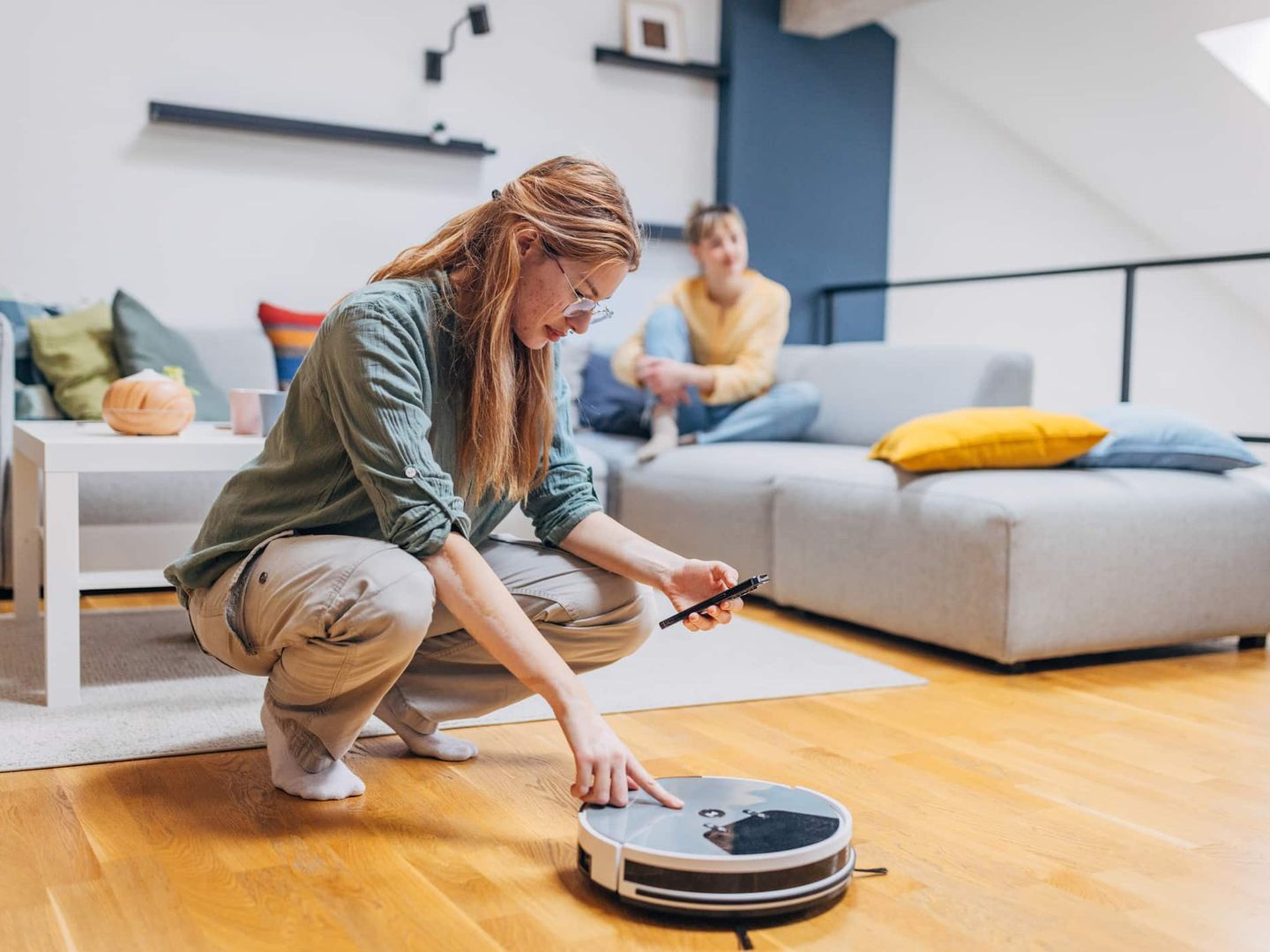 Frau programmiert Staubsaugerroboter per Smartphone, Partner sitzt im Hintergrund Getty