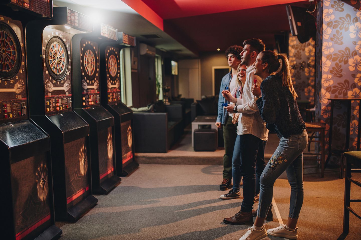 Vier junge Erwachsene spielen gemeinsam Dart an elektronischen Dartscheiben in einer Bar oder Spielhalle.