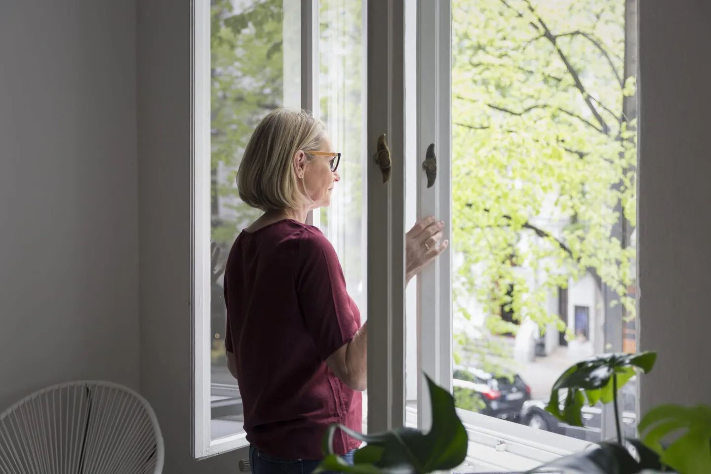 Frau schaut nachdenklich aus einem geöffneten Fenster auf eine grüne Baumallee.
