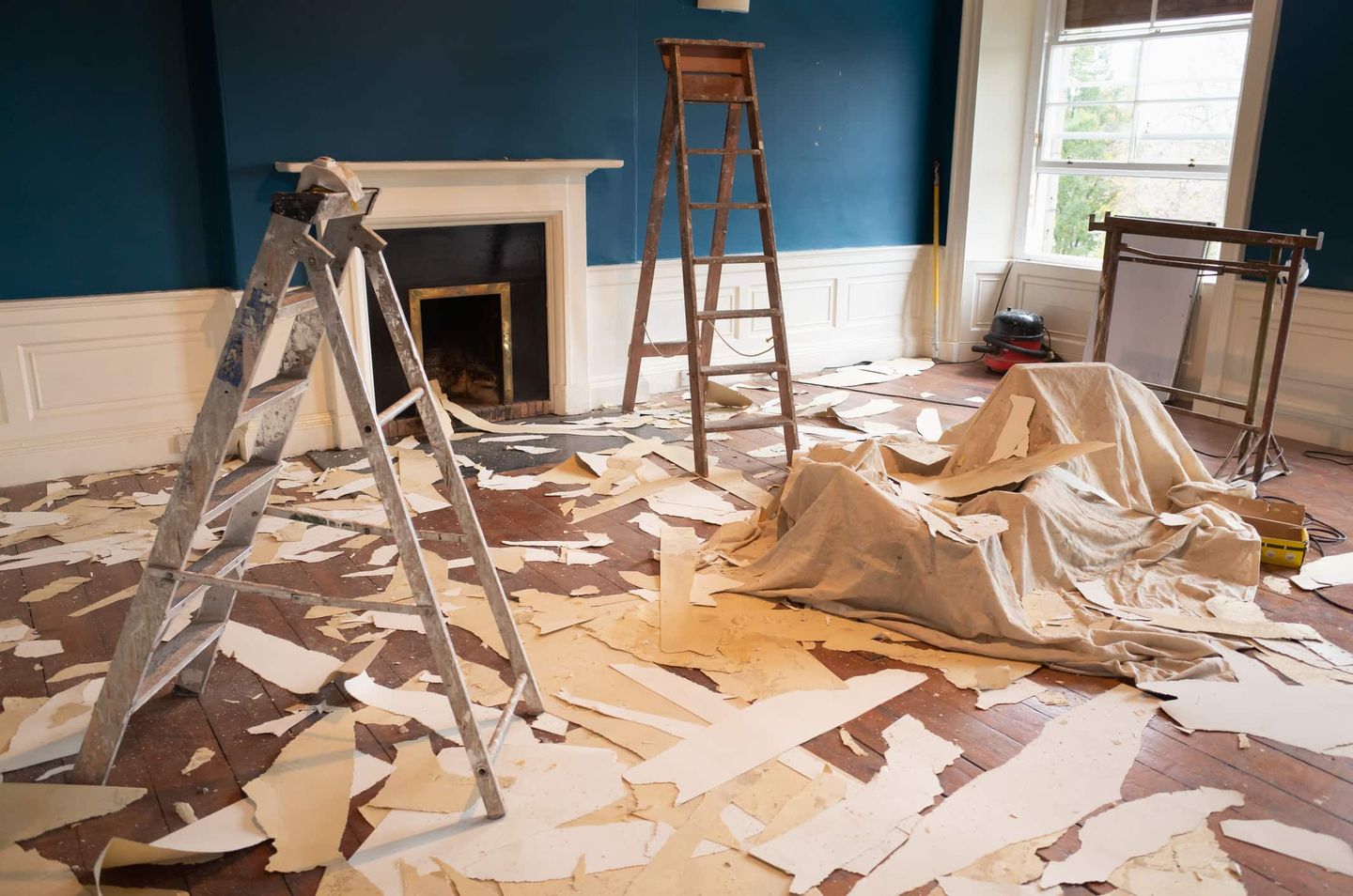Ein Wohnzimmer mit abgerissener Tapete, Leitern und Abdeckplanen während einer Renovierung. Getty