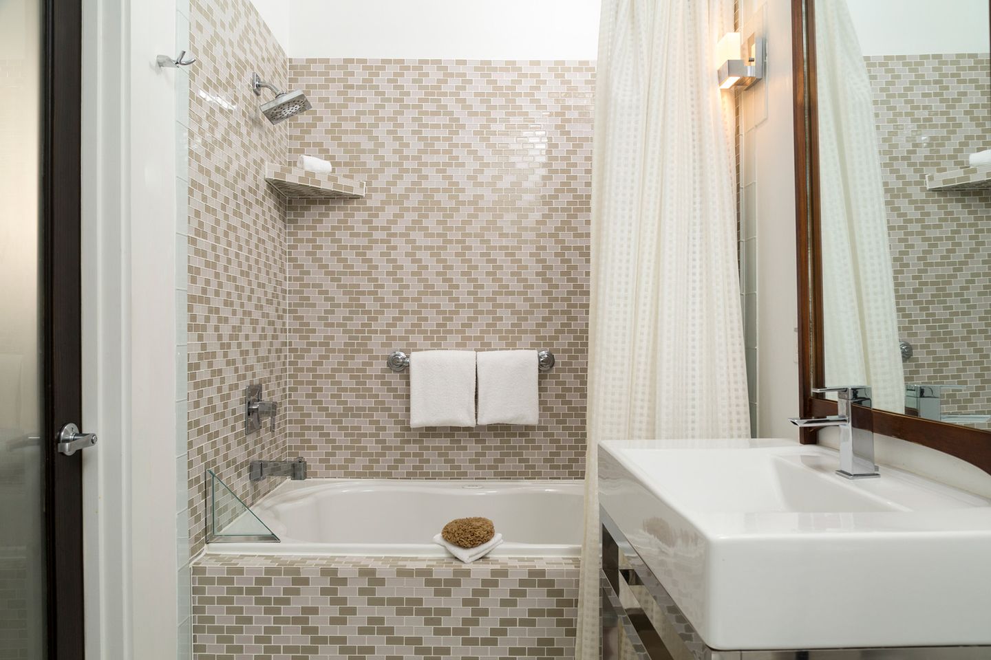 Ein helles Badezimmer mit beige-weißem Mosaikfliesenmuster, Badewanne mit Duschkopf, Duschvorhang und weißen Handtüchern.