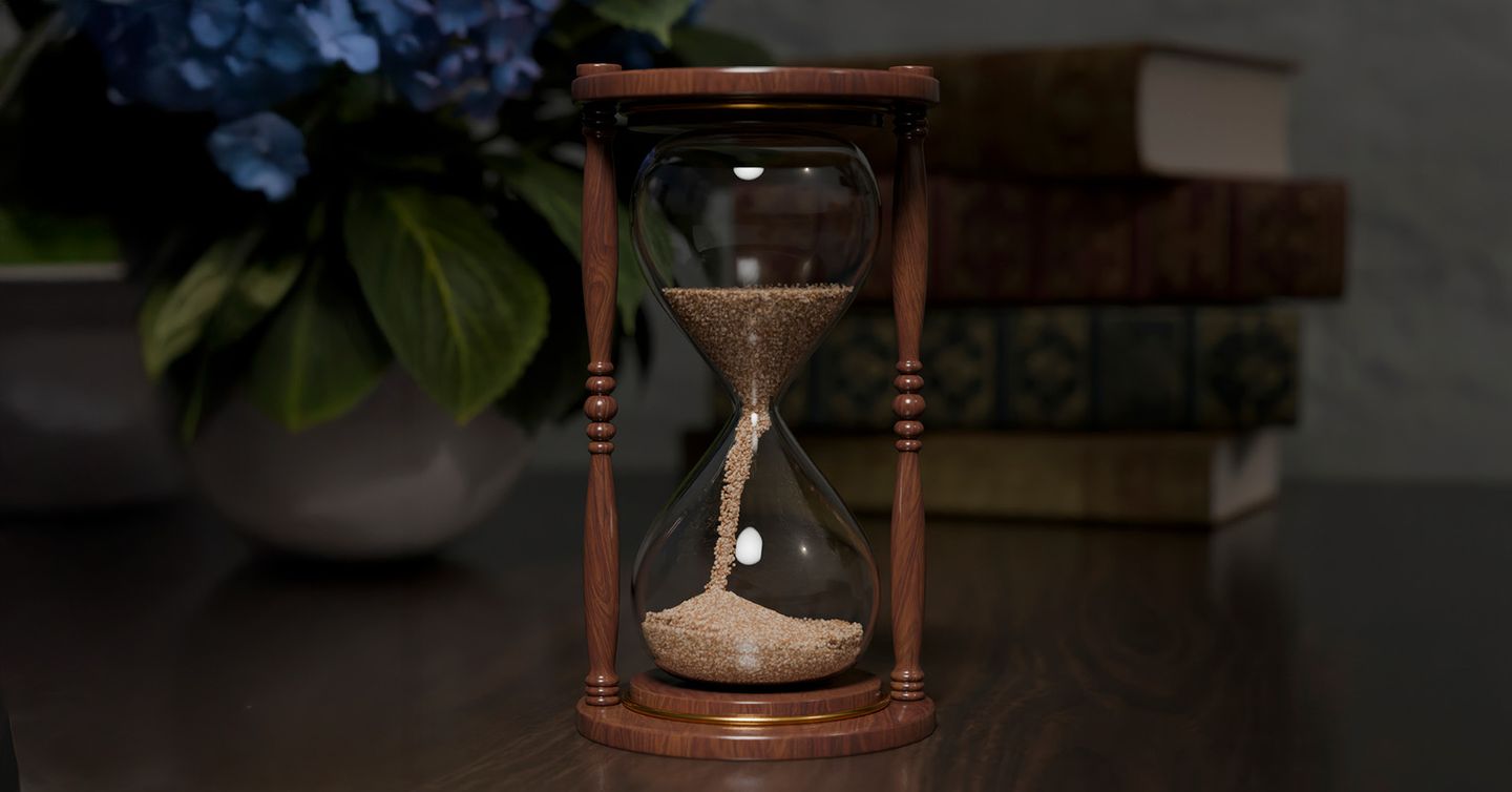 Eine klassische Sanduhr mit Holzrahmen steht auf einem Tisch. Der Sand rieselt von der oberen Kammer in die untere. Im Hintergrund sind unscharf Bücher und eine Vase mit blauen Blüten zu sehen.