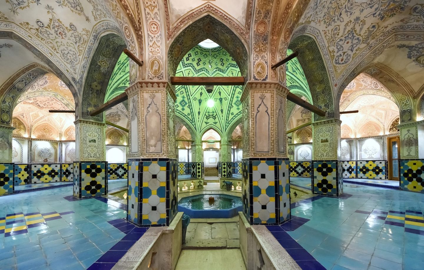 Innenansicht eines historischen Hammams mit reich verzierten, farbenfrohen Mosaiken, hohen Gewölbedecken und einem zentralen Brunnen unter grünen Kuppeln