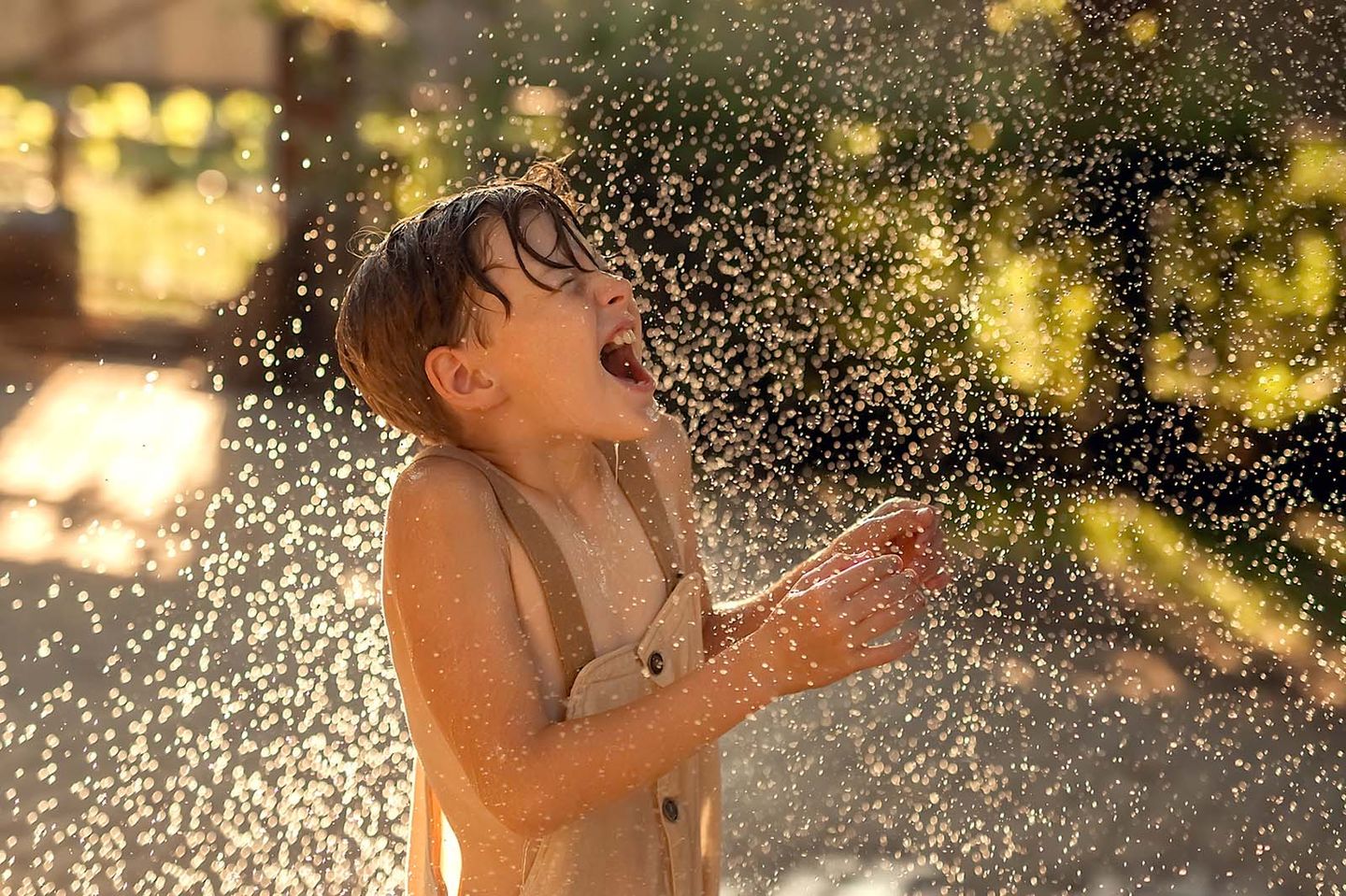 Ein lachendes Kind spielt im Freien mit Wasser. Das Wasser spritzt in feinen Tropfen in die Luft, während das Kind, durchnässt und mit geschlossenen Augen, den Moment sichtlich genießt.
