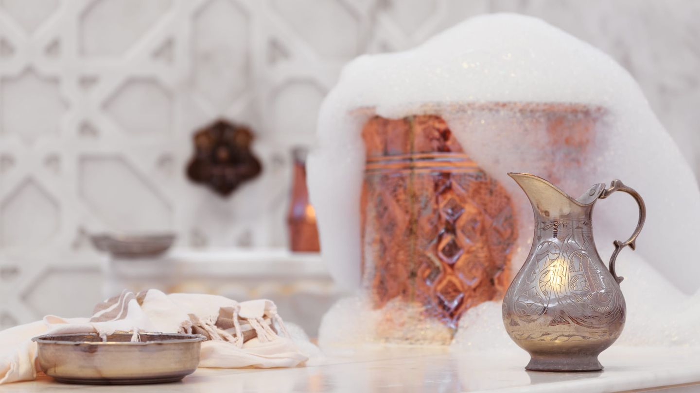 Detailaufnahme eines traditionellen Hammam-Zubehörs mit einem Kupferkessel, umgeben von Seifenschaum, und einem kunstvoll verzierten Wasserkrug im Vordergrund