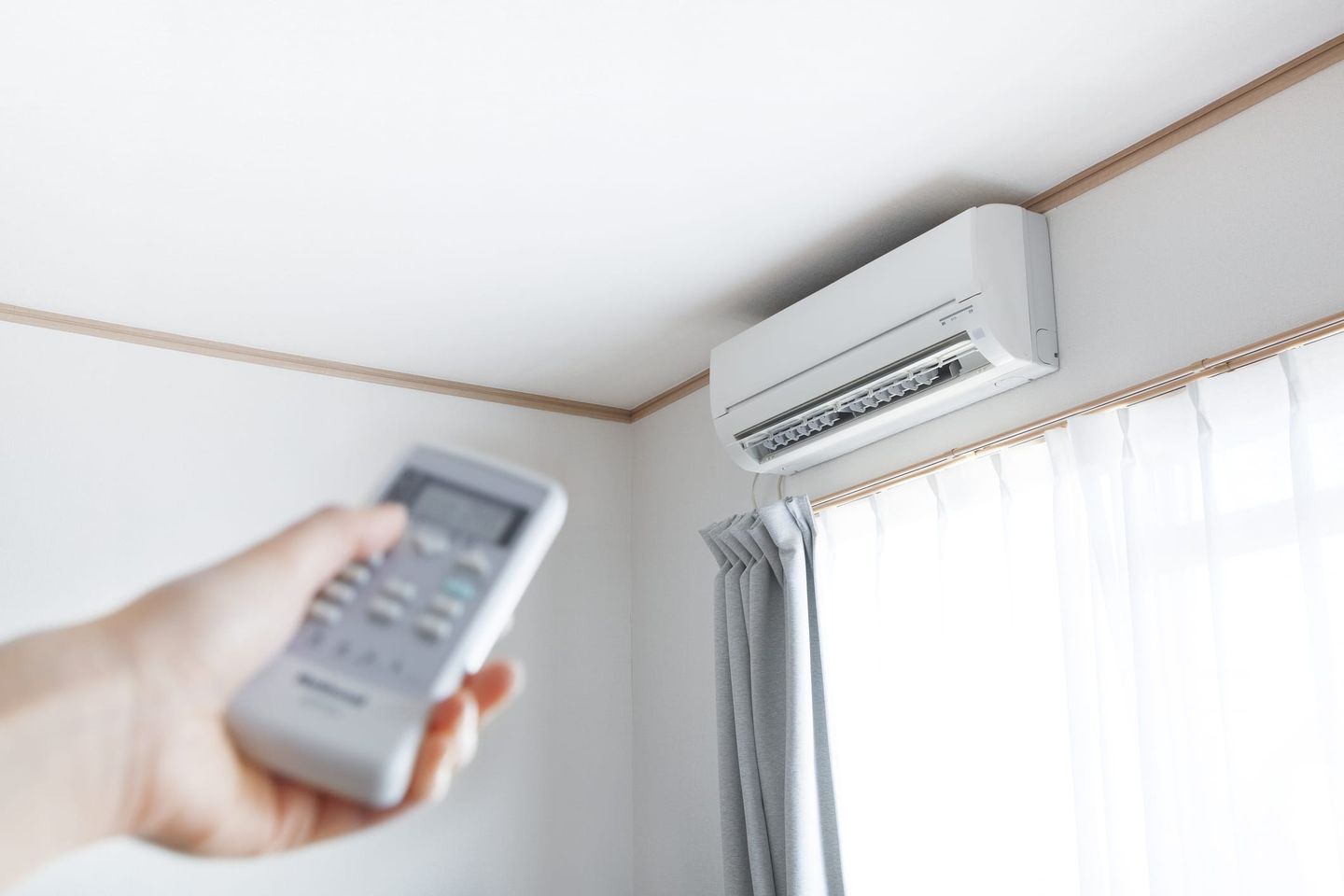 Nahaufnahme einer Hand mit Fernbedienung, die auf eine an der Wand montierte Klimaanlage im hellen Raum gerichtet ist.