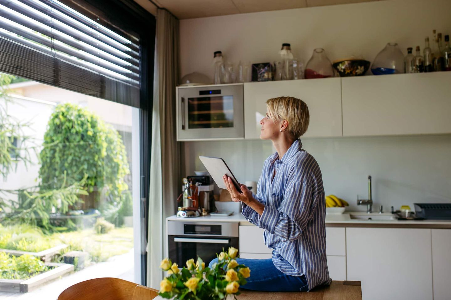 Frau sitzt in moderner Küche mit Tablet und steuert Smart-Home-Funktionen. Getty