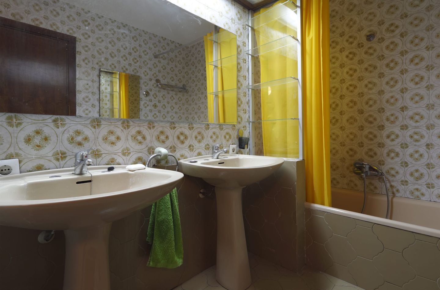 Badezimmer im Retro-Stil mit zwei beigen Waschbecken auf Sockeln, gemusterten Fliesen in Grün- und Brauntönen, gelbem Duschvorhang und großem Spiegel.