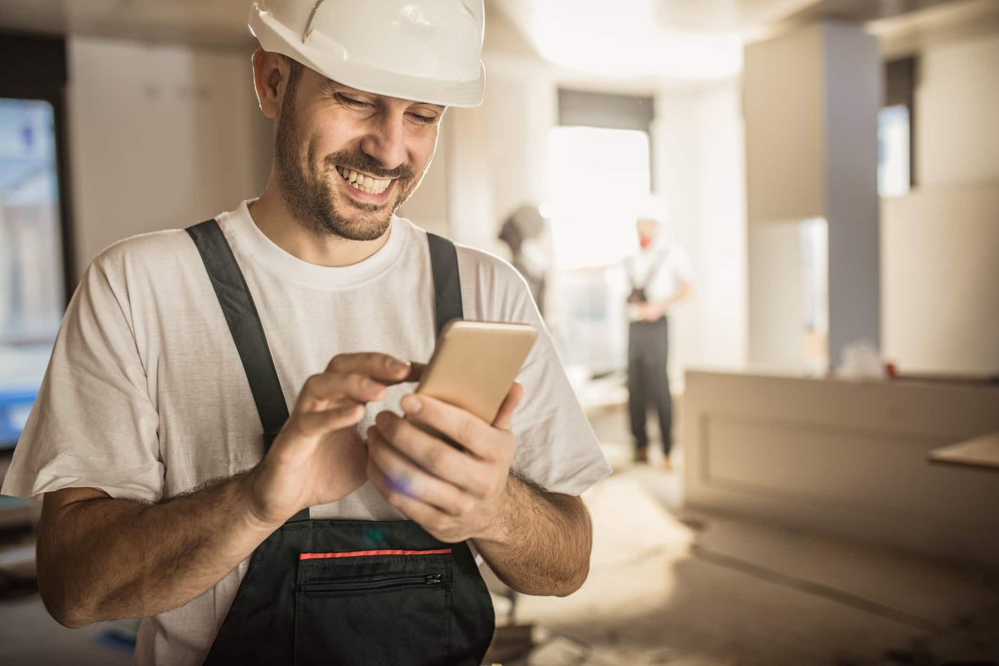 Handwerker mit Bauhelm und Latzhose schaut lächelnd auf sein Smartphone während einer Renovierung. Getty