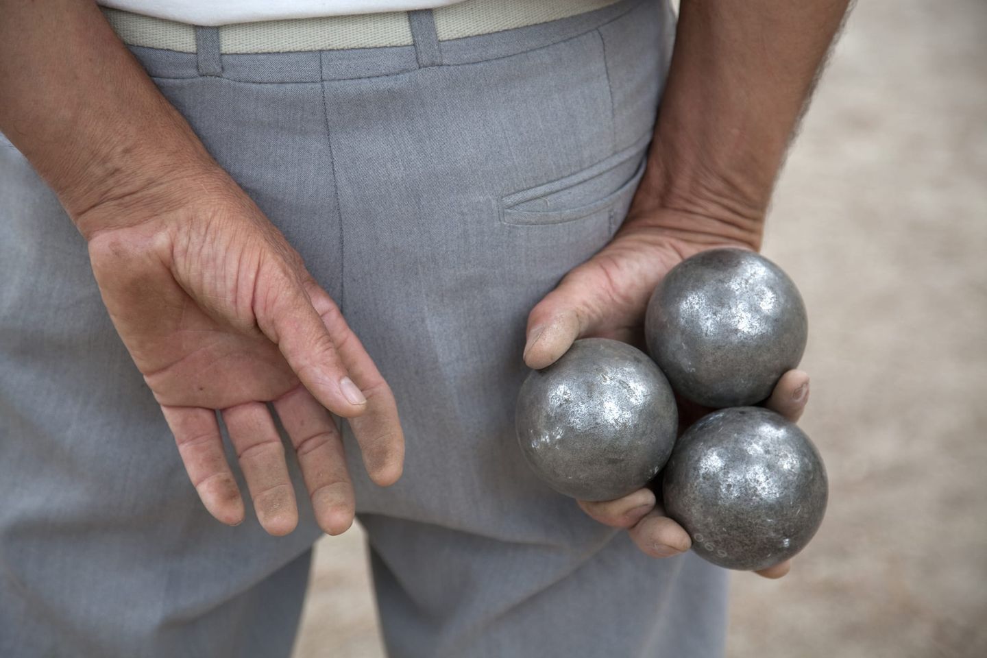 Nahaufnahme eines Mannes in grauer Hose, der hinter seinem Rücken drei silberne Boule-Kugeln in einer Hand hält.