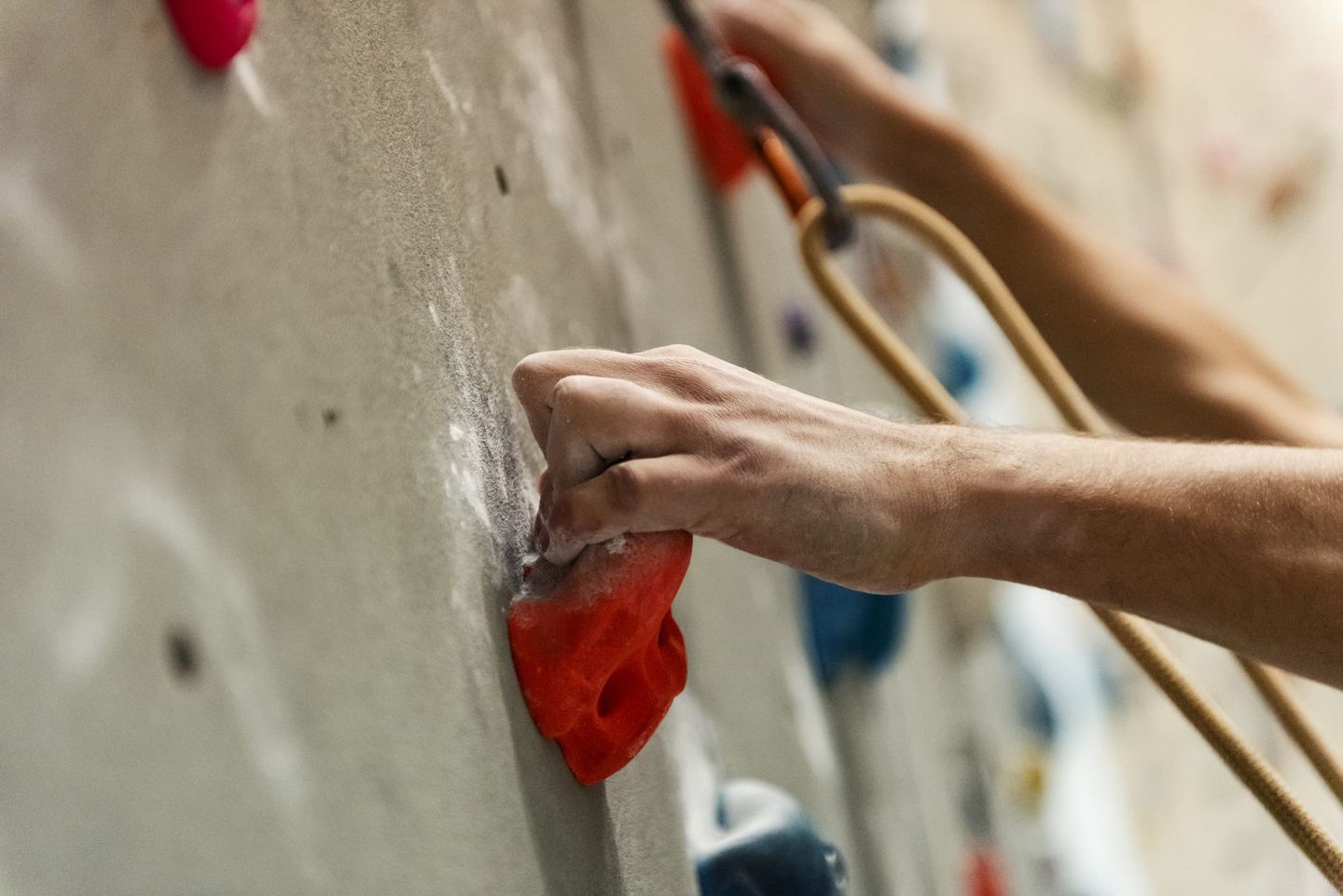 Nahaufnahme einer Hand, die einen roten Klettergriff an einer Kletterwand umklammert, während ein Seil zur Sicherung sichtbar ist.