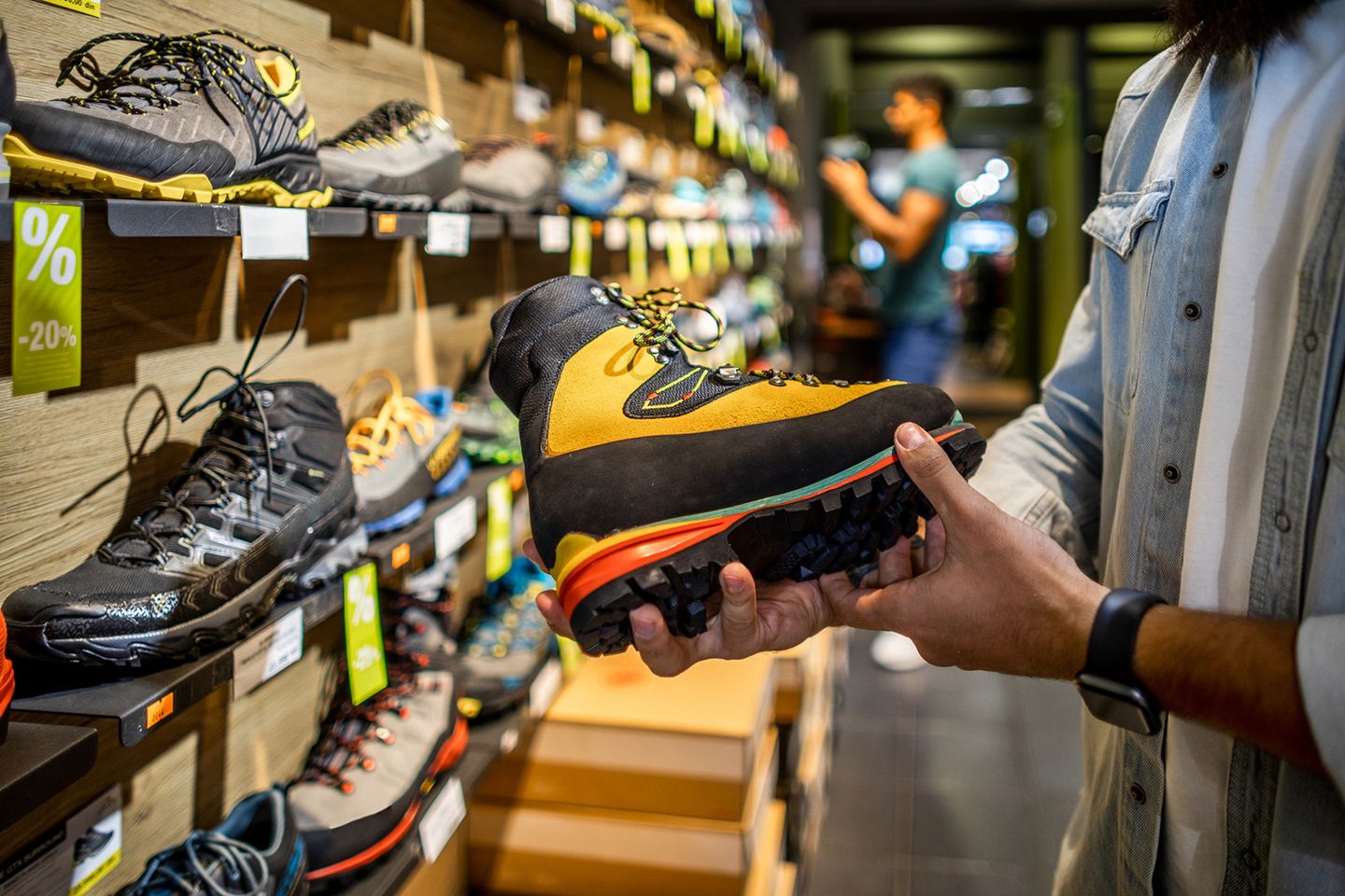 Eine Person hält einen gelb-schwarzen Wanderschuh mit robuster Profilsohle in den Händen, während sie sich in einem Schuhgeschäft befindet. Im Hintergrund sind weitere Sportschuhe auf Regalen und ein anderer Kunde unscharf zu sehen.