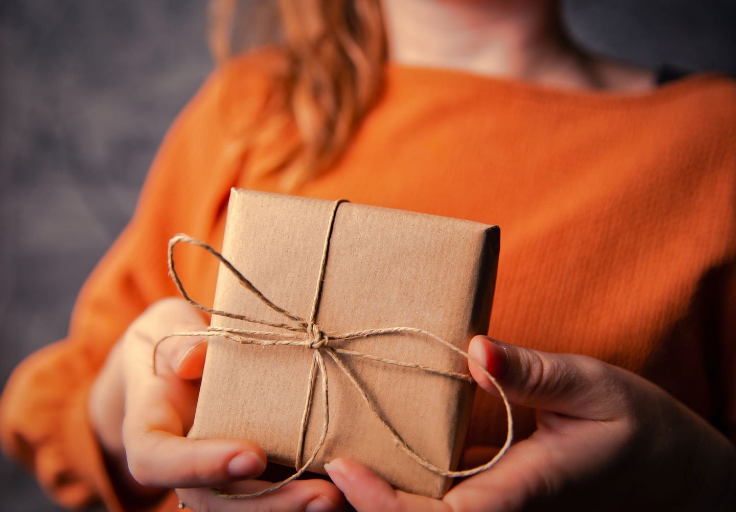 Nahaufnahme von zwei Händen, die ein kleines Geschenk in braunem Packpapier mit einer einfachen Kordel-Schleife halten, vor einem Hintergrund in orangefarbener Kleidung