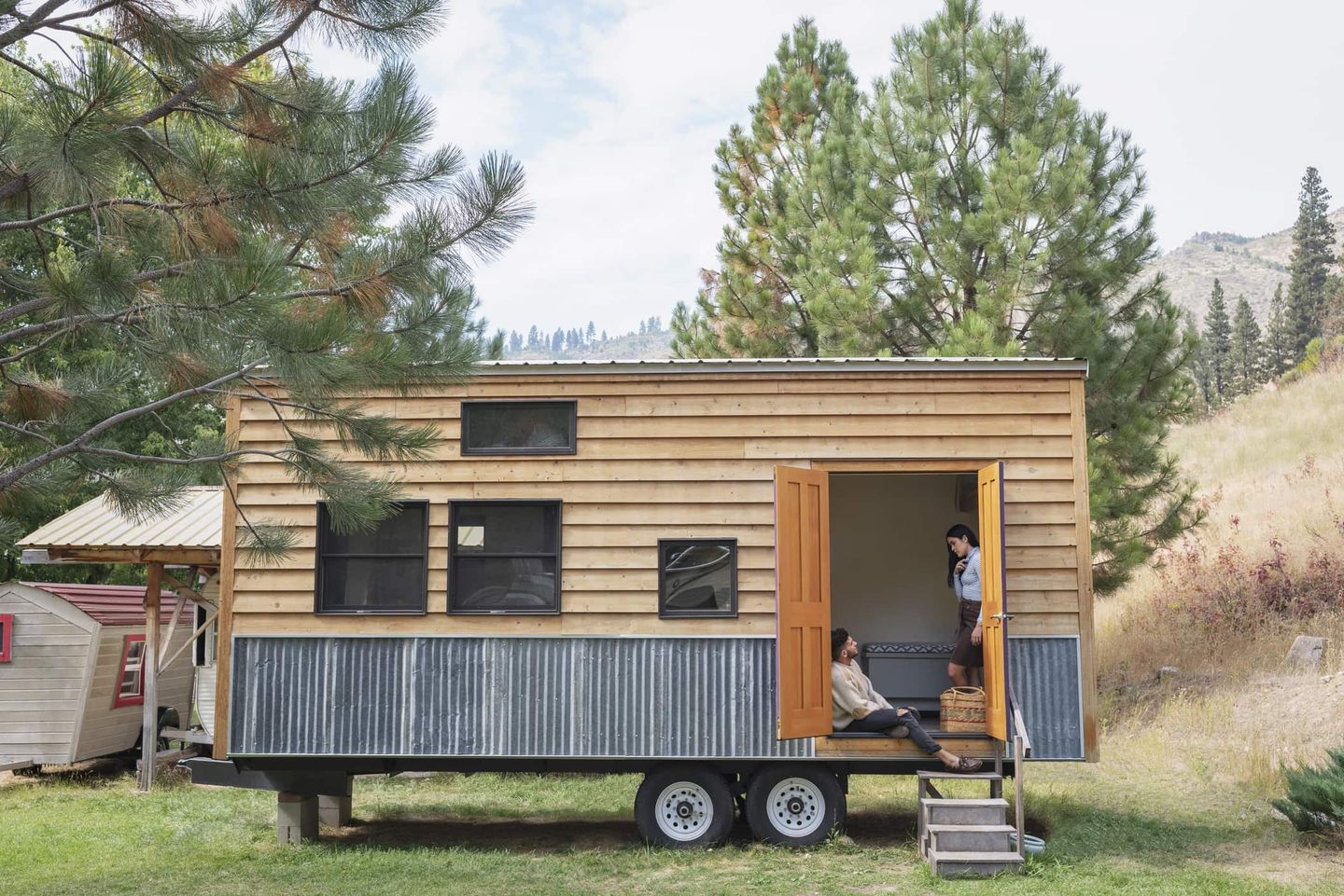 Tiny House auf Rädern mit Holzfassade im Grünen, zwei Personen sitzen im Eingang Getty