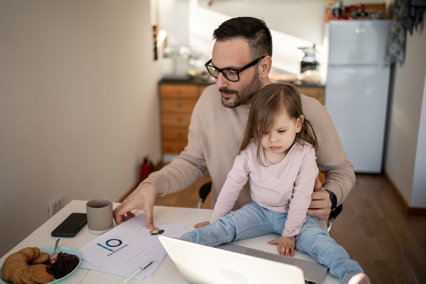 Vater arbeitet zu Hause am Laptop, während sein Kind auf dem Schreibtisch sitzt Mirjana Pusicic via Getty Images