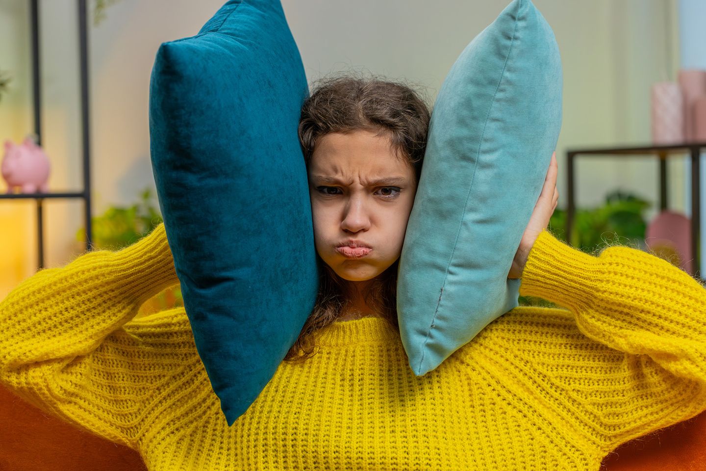 Ein genervtes Kind in gelbem Pullover drückt zwei Kissen gegen seinen Kopf und bläst die Wangen auf, als wolle es Lärm ausblenden.