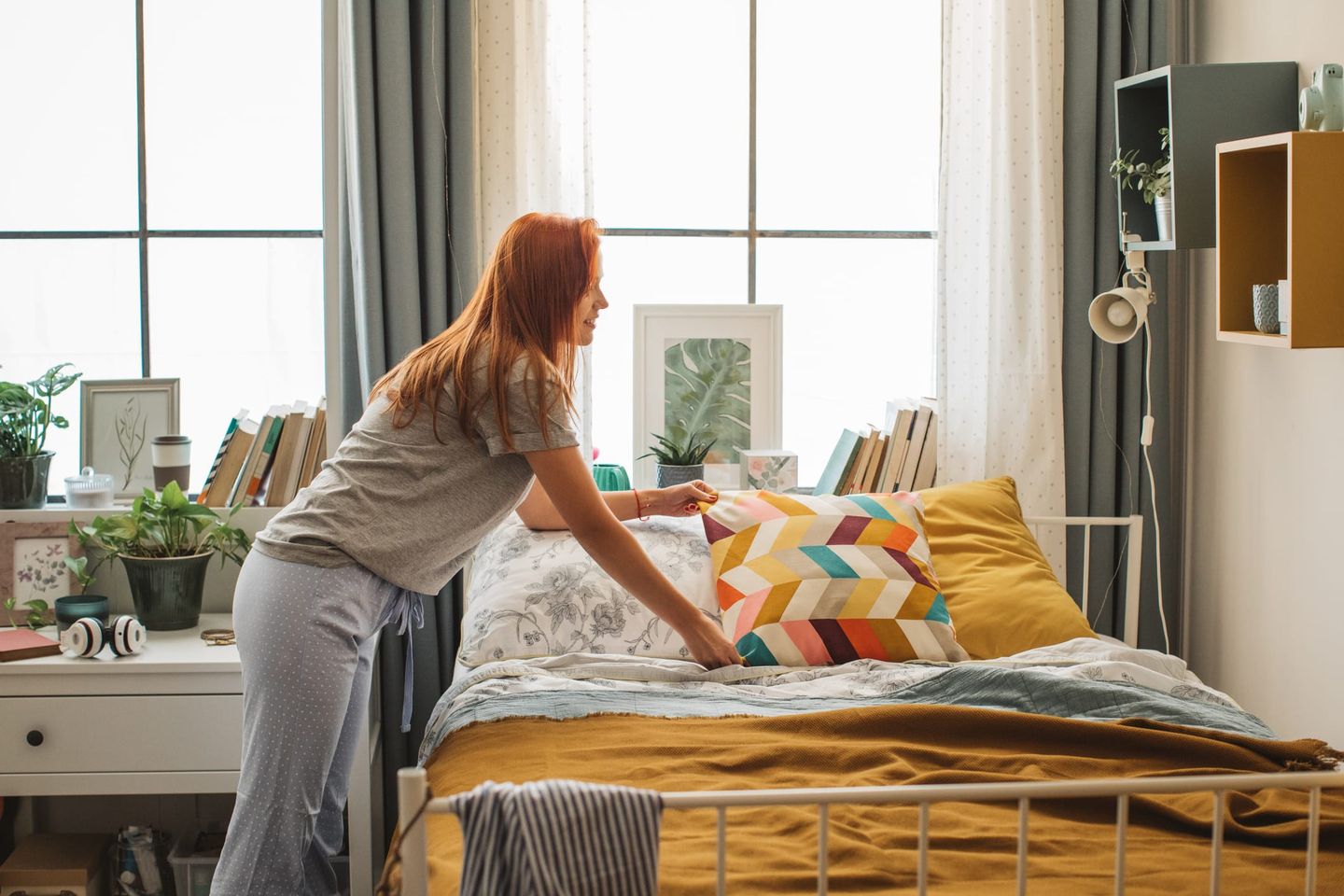 Eine Frau mit roten Haaren richtet ein buntes Zierkissen auf einem Bett in einem hellen, gemütlichen Schlafzimmer mit Pflanzen, Büchern und Dekorationen.