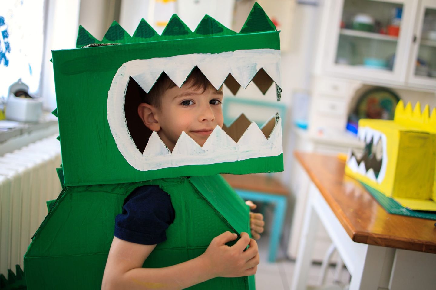 Ein Junge im selbst gebastelten grünen Dinosaurier-Kostüm aus Karton feiert fröhlich Karneval zu Hause